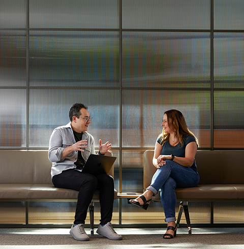 Dois colegas conversam sentados em bancos em uma área comum.