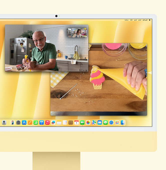 iMac باللون الأصفر. عرض توضيحي لميزة العرض الرأسي. شاشة تعرض صانعة حلويات تشارك في مكالمة فيديو مع شخص آخر. وتعرض إحدى الشاشتين صانعة الحلويات ووجه الرجل، بينما تعرض الشاشة الأخرى يدي صانعة الحلويات وهي تزيّن قطعة بسكويت