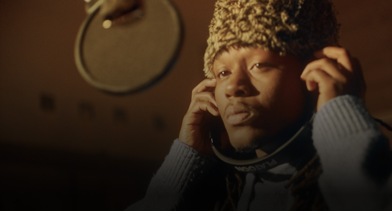 Foto de Kiddo K en un estudio de música de pie delante de un micrófono y con unos auriculares puestos