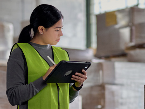 Debout dans une usine, une femme effectue le suivi des stocks sur un iPad. Derrière elle se trouvent plusieurs piles de grandes boîtes.
