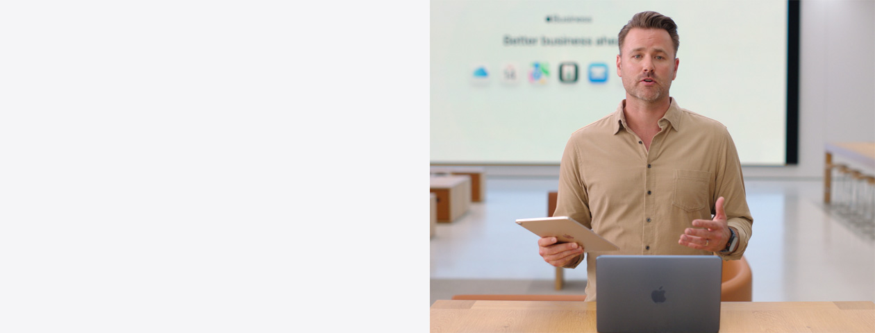 発表者がiPadを手に持ち、テーブルの上に置いたMacBookの前に立って話している。大型スクリーンにはビジネスで使う様々なアプリのアイコンが表示されている