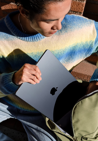Seorang mahasiswa sedang duduk di luar. Dia mengambil MacBook dari tas ranselnya.