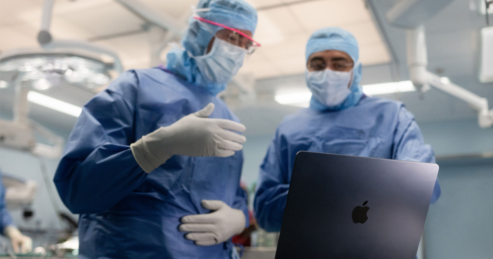 手術室でMacBookを見ている2人の外科医。1人が手でジェスチャーし、もう1人はそれを見ている