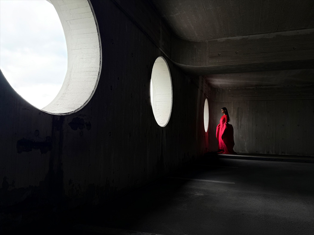 A woman wearing a long red dress stands in an empty room being photographed. The Photo app toggles through the following focal lengths to demonstrate the capabilities of the pro camera system: 200 mm, 100 mm, 48 mm, 35 mm, 28 mm, 13 mm and macro
