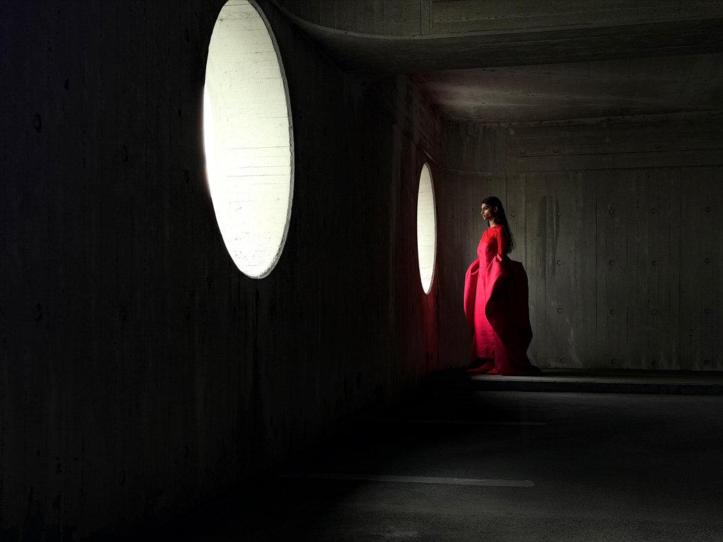 A woman wearing a long red dress stands in an empty room being photographed. The Photo app toggles through the following focal lengths to demonstrate the capabilities of the pro camera system: 200 mm, 100 mm, 48 mm, 35 mm, 28 mm, 13 mm and macro