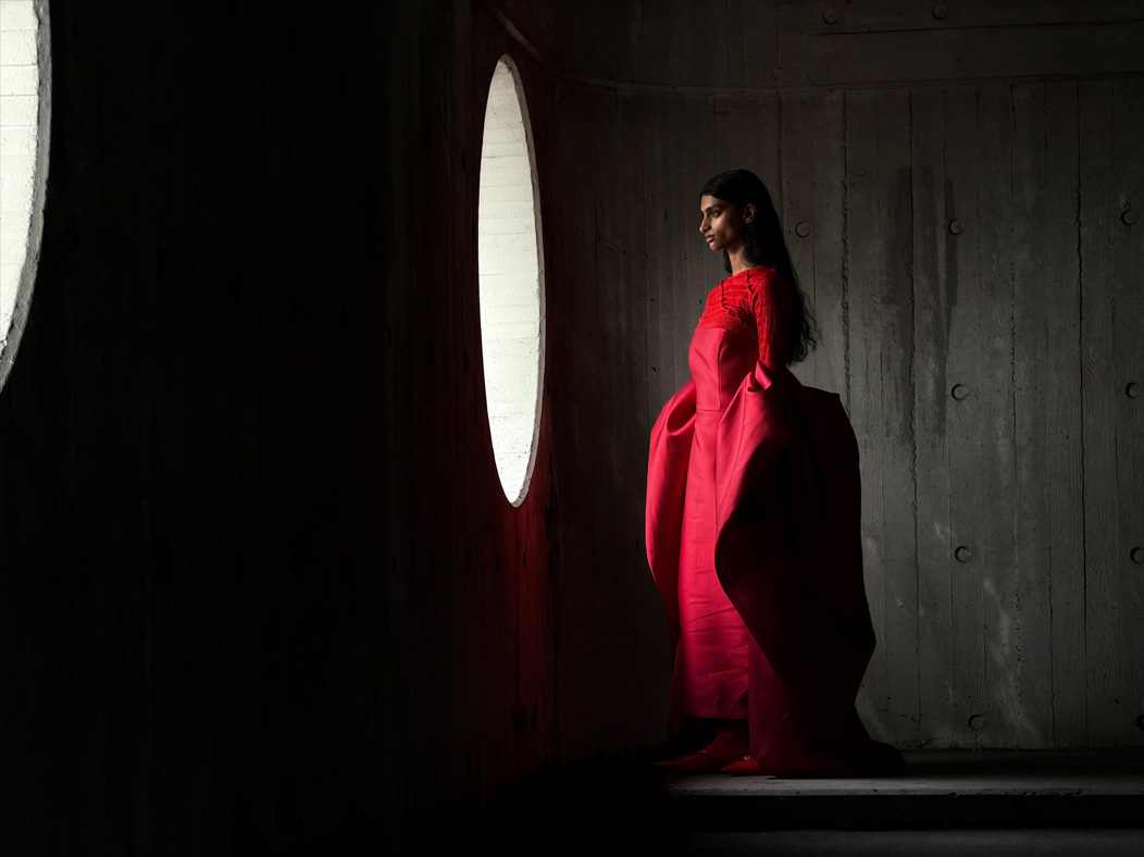 A woman wearing a long red dress stands in an empty room being photographed. The Photo app toggles through the focal lengths to demonstrate the capabilities of the pro camera system, showcasing: 100 mm