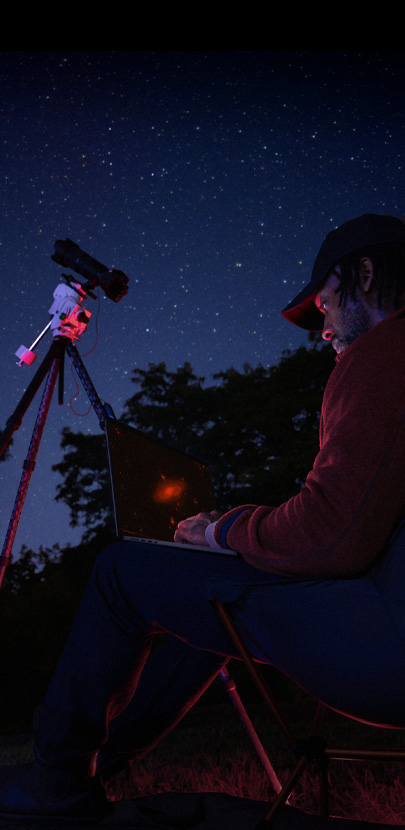 A person outside at night using a MacBook Pro on their lap, connected to a device on a tripod pointed towards the starry sky.
