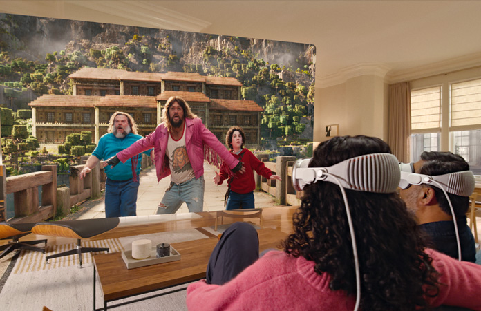 Two people both wearing Apple Vision Pro sit side by side on a couch watching: A Minecraft Movie, displayed on a screen stretching the across room