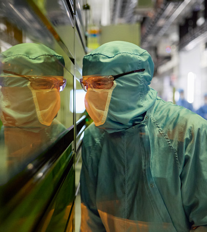 フルカバーの作業服を着た1人の従業員が半導体製造装置の窓から中を覗いている